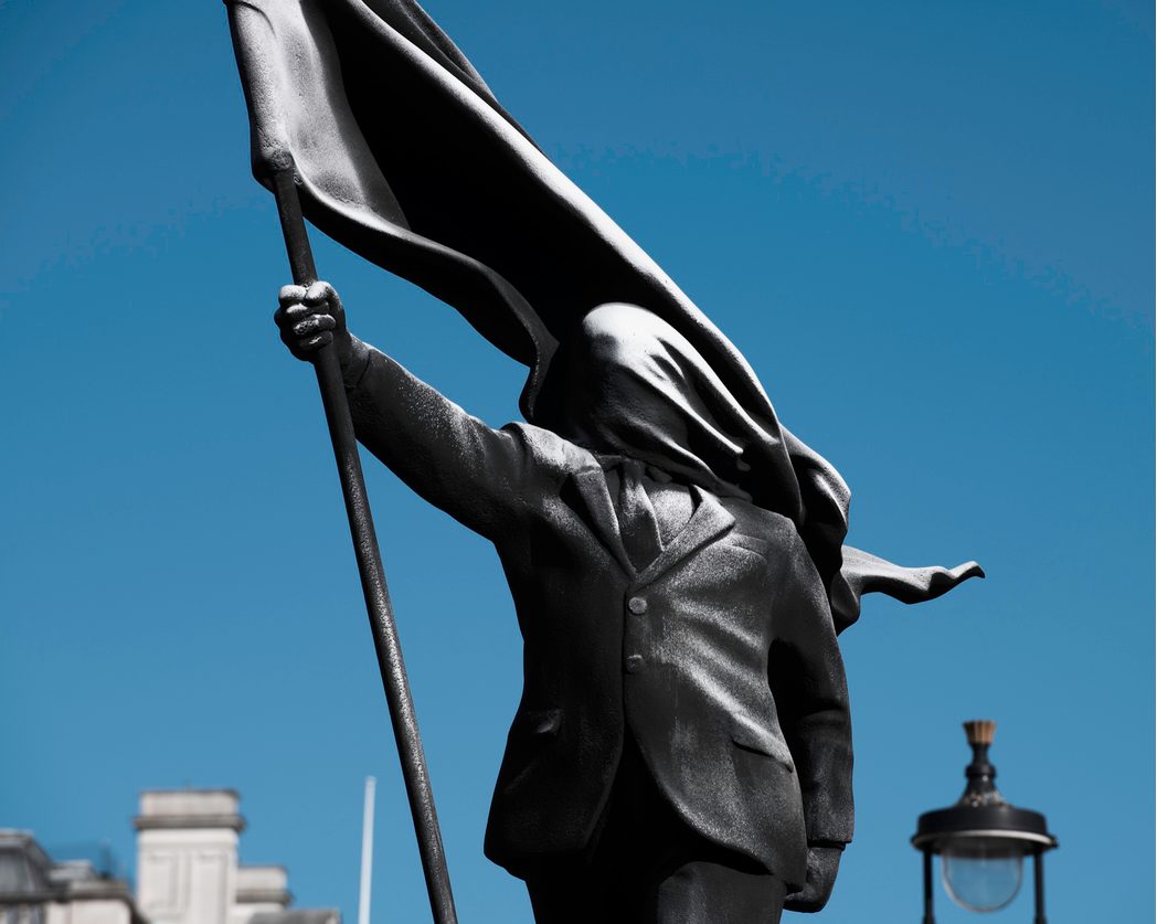 Statue sombre d'un homme en costume marchant en avant, brandissant un drapeau dont l'étoffe lui recouvre le visage, sur fond de ciel bleu.